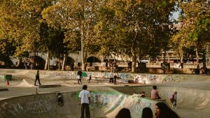 Teenagers skateboarding and hanging out at a graffiti-covered outdoor skate park, with tall trees and a stone bridge in the background.