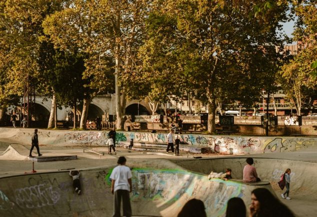 Teenagers skateboarding and hanging out at a graffiti-covered outdoor skate park, with tall trees and a stone bridge in the background.